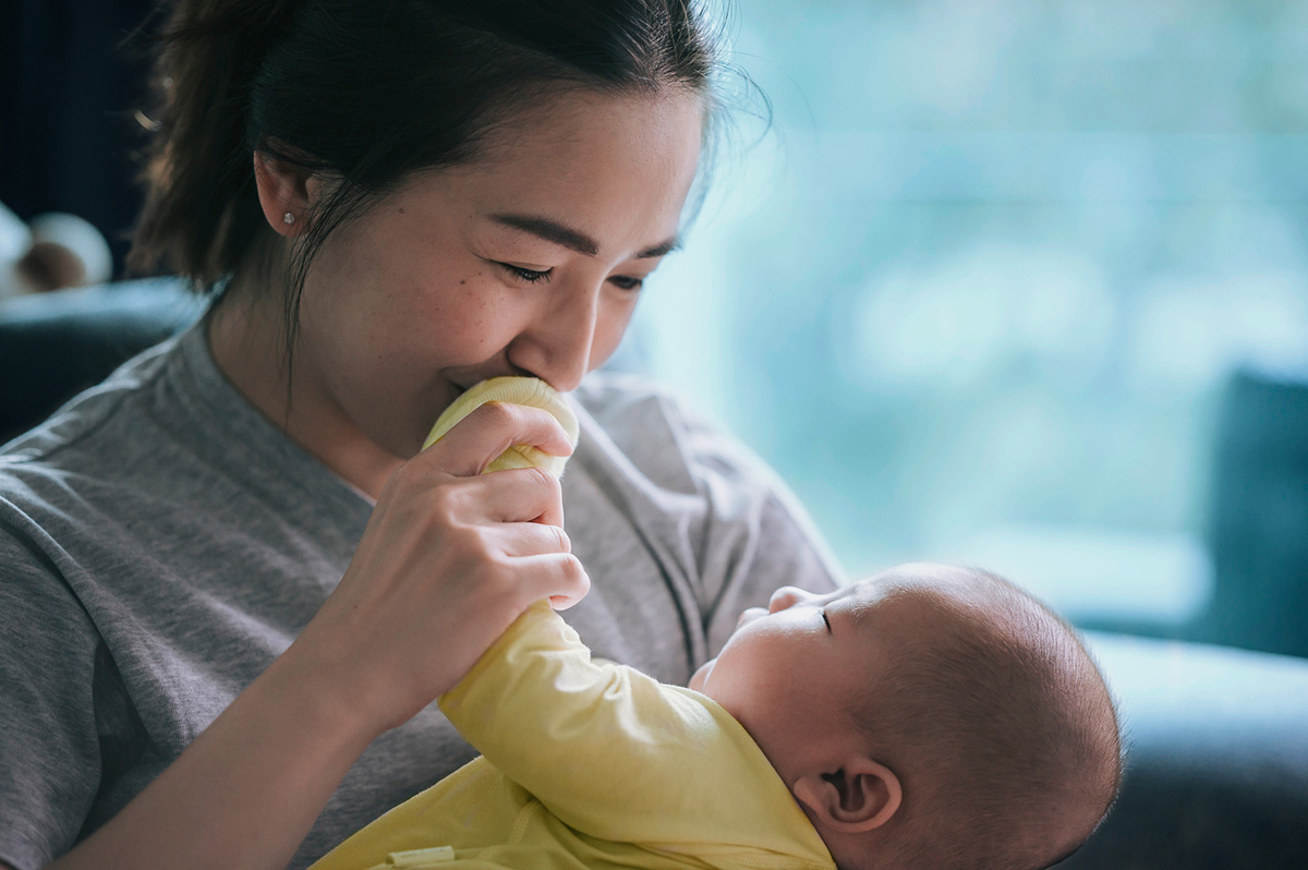 A smiling mother holds her young baby and kisses the baby’s hand.