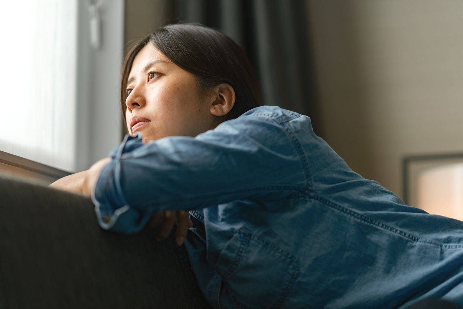 A young woman inside her home looks out her window longingly.