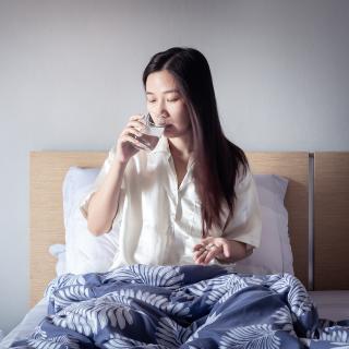 A young woman takes a pill with a glass of water from her bed.